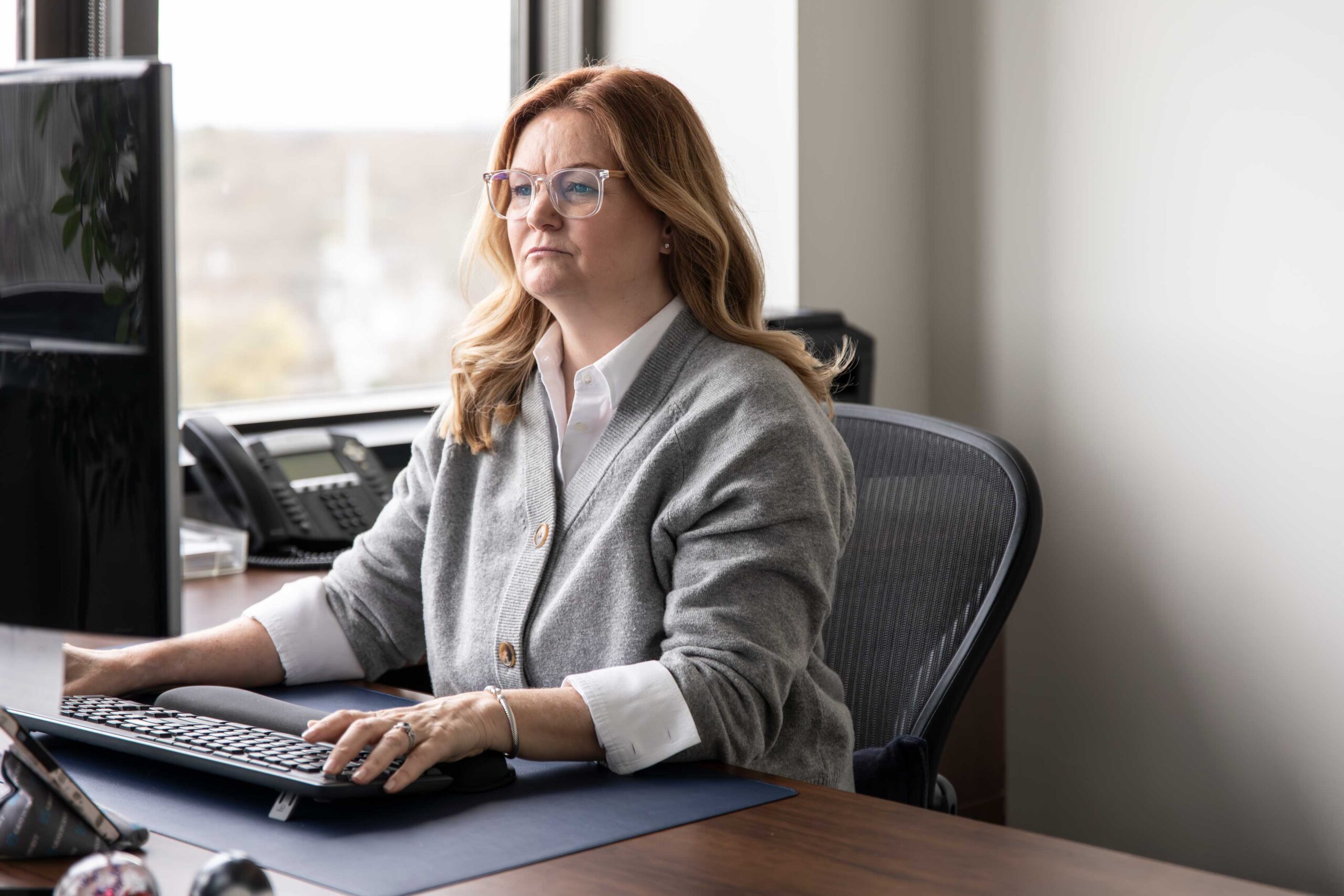 Renee has strawberry blonde hair and is wearing a gray cardigan over a white blouse while she sits at her computer.